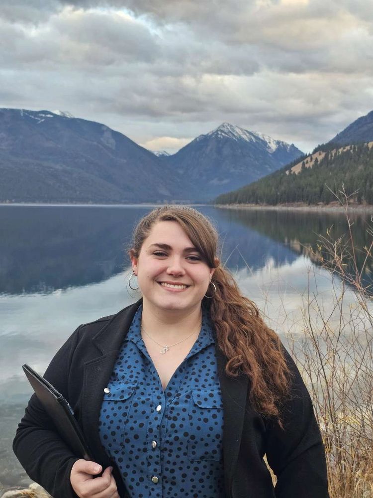 Smiling woman standing in front of a serene lake with mountains.