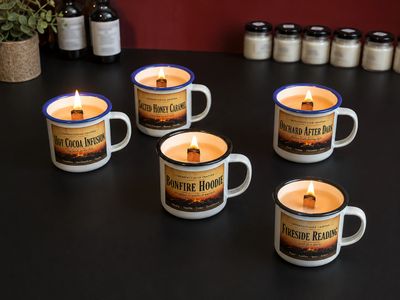Five lit candles in mugs with cozy labels on a black surface.