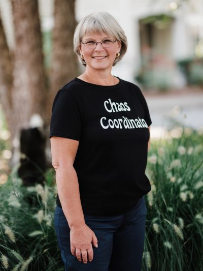 Smiling woman wearing glasses and "Chaos Coordinator" t-shirt outdoors.