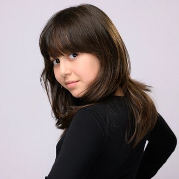 Young girl with brown hair and bangs wearing a black top, looking over her shoulder.