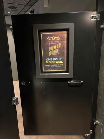 Black restroom stall door with a neon sign advertising Power Hour drinks.
