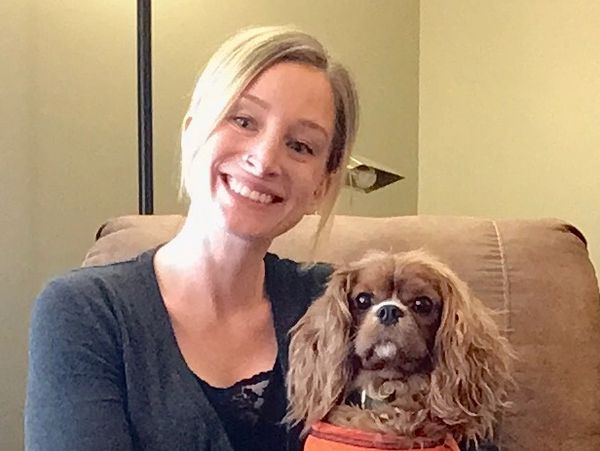 Smiling woman sitting on a couch with a small dog wearing an orange shirt.