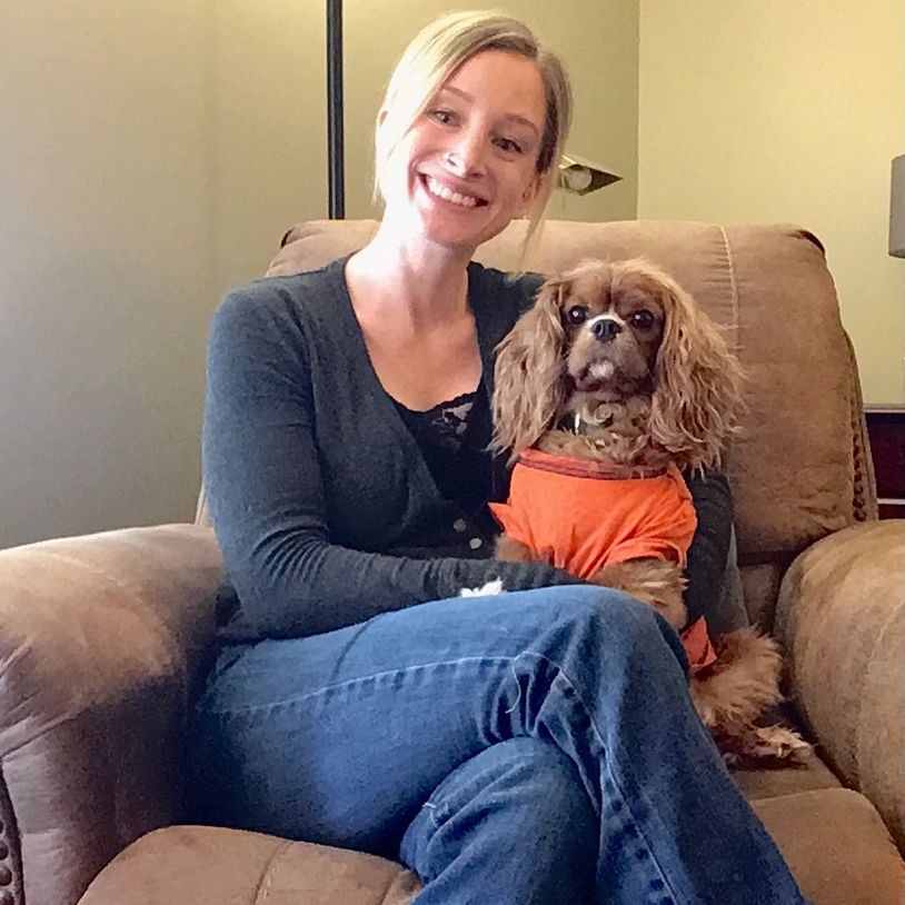 Smiling woman sitting on a couch with a small dog wearing an orange shirt.