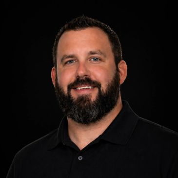 Smiling man with beard in black polo against black background.