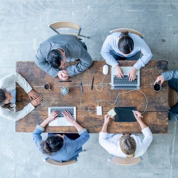 Top view of six diverse professionals collaborating around a wooden table with laptops and coffee.