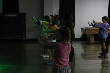 Children practicing dance moves with outstretched arms in a dimly lit room.