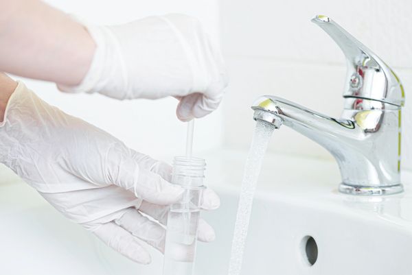 Gloved hands collecting a water sample from a running faucet into a small vial.