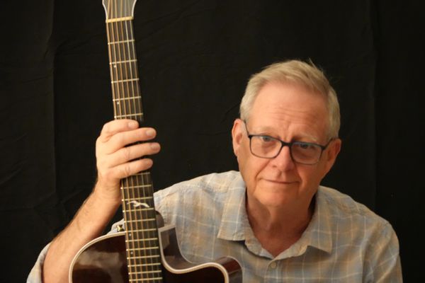 Older man holding guitar with black backdrop.