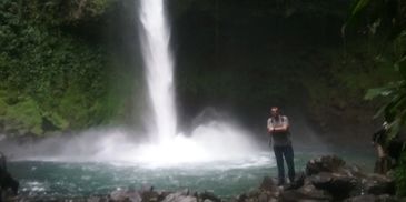 Person standing near a powerful waterfall in a lush green forest.
