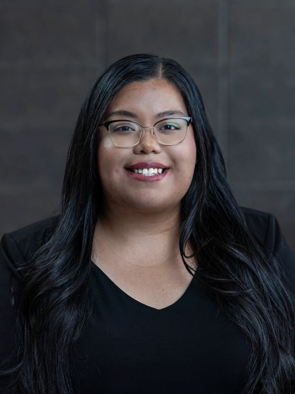 Portrait of a smiling woman with long black hair and glasses against a dark background.