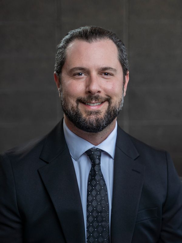 Smiling man in a suit and tie with a beard against a dark background.