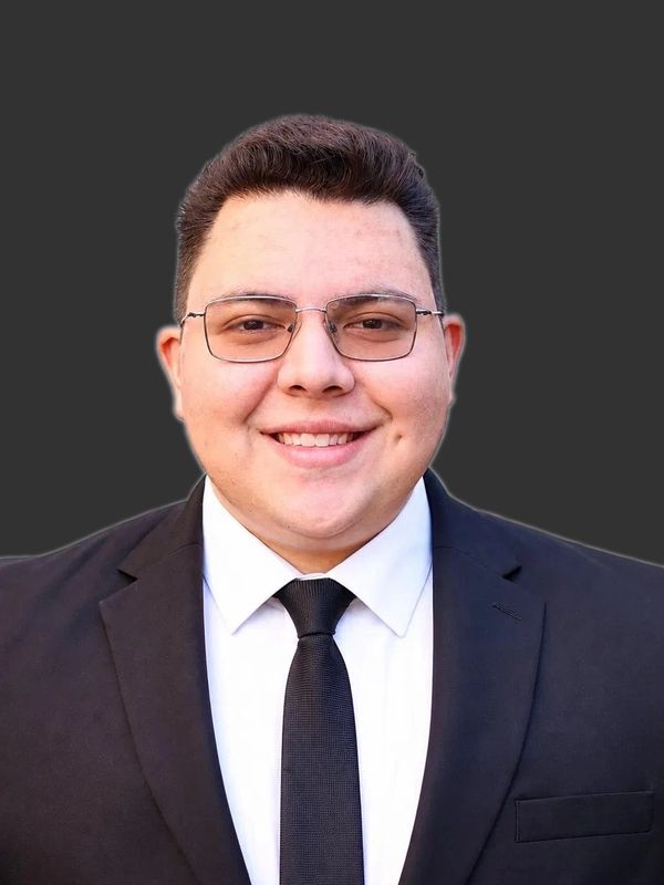 Young man in a black suit and glasses smiling against a dark background.