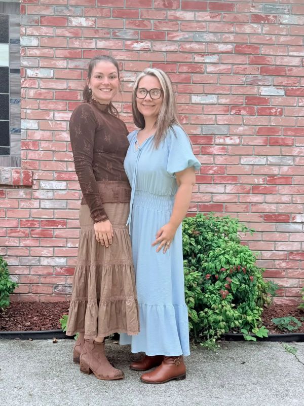 Two women posing in front of a brick wall with plants.