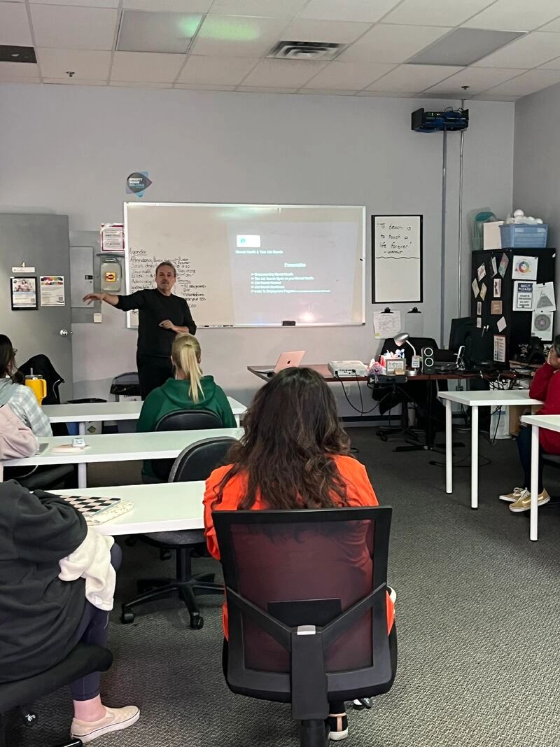 A teacher presents to students in a classroom with a projected slide on a whiteboard.