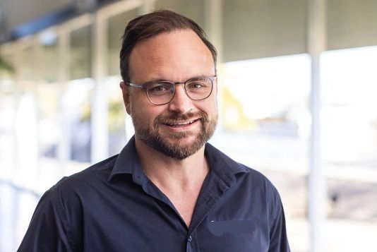 Man with glasses and beard smiling, wearing a dark shirt indoors.