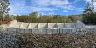 Concrete dam structure surrounded by rocky terrain and trees under a blue sky.