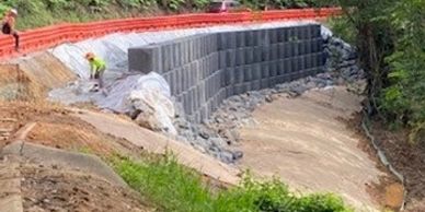 Workers building a retaining wall with concrete blocks near a forested area.