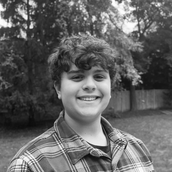 Smiling young person outdoors in a plaid shirt with trees in the background.