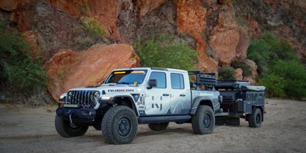 White Jeep Rubicon towing an off-road camping trailer in a rocky desert landscape.