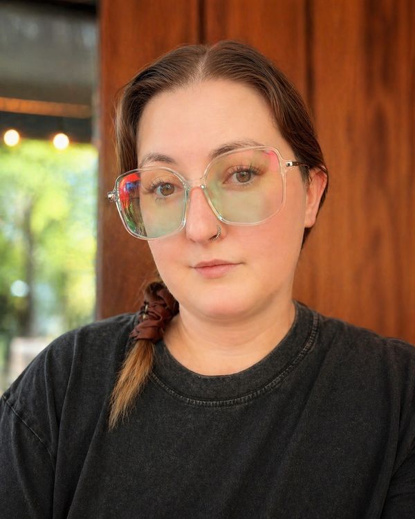 Person wearing oversized clear glasses and a black shirt with a wooden background.