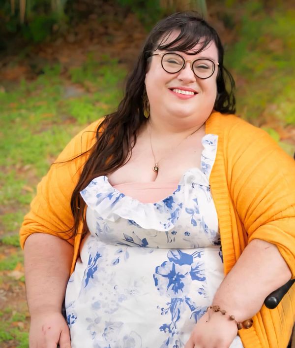Smiling woman in glasses wearing a floral dress and yellow cardigan outdoors.