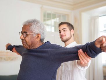 A young man assists an elderly man with arm stretching exercises indoors.