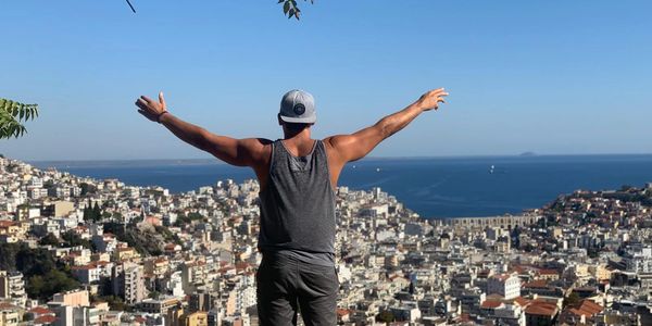 Man with arms outstretched overlooking a coastal city and sea on a clear day.