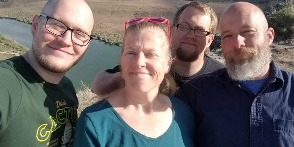 Four smiling people taking a selfie outdoors by a river under sunny weather.