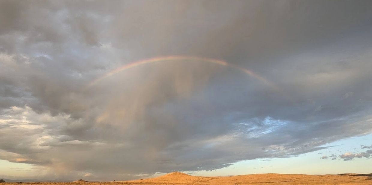 A faint rainbow arches over a dry, golden landscape under a cloudy sky.