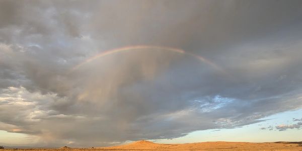 A faint rainbow arches over a dry, desert landscape under a cloudy sky.