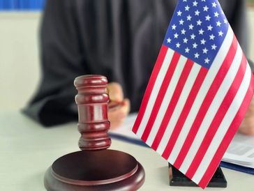 A judge's gavel and a small American flag on a desk, with a person in the background.