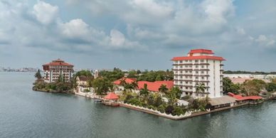 A waterfront complex with red-roofed buildings surrounded by calm water under a cloudy sky.