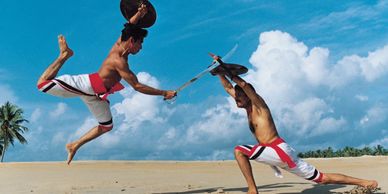 Two men in traditional attire engage in a dynamic martial arts duel on a sandy beach.