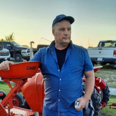 Man in blue shirt standing near red tractor at sunset.