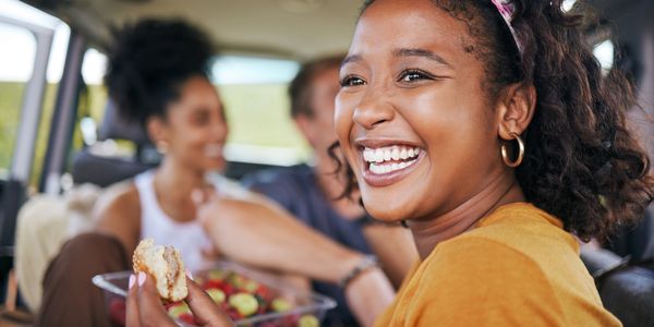 A cheerful young woman enjoys a fresh, healthy snack while laughing with friends on a road trip.