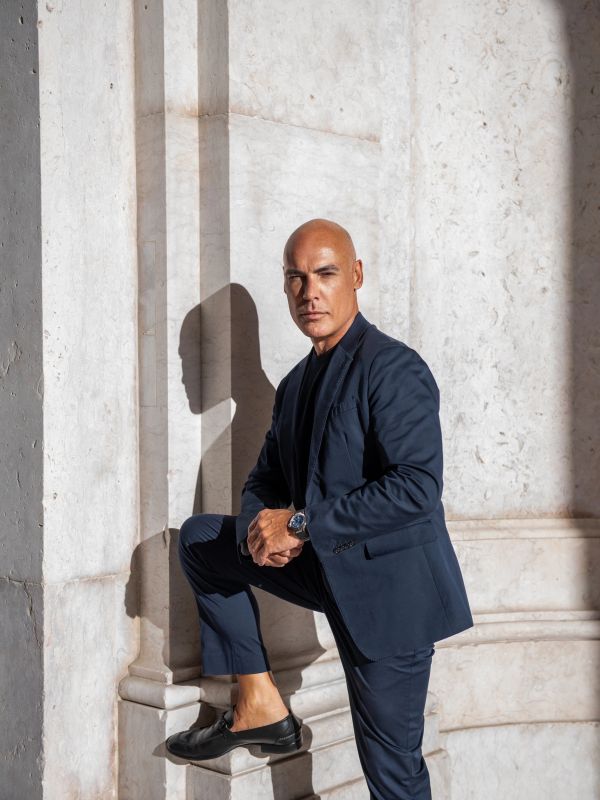 Confident man in a navy suit poses against a marble wall with sunlight casting his shadow.