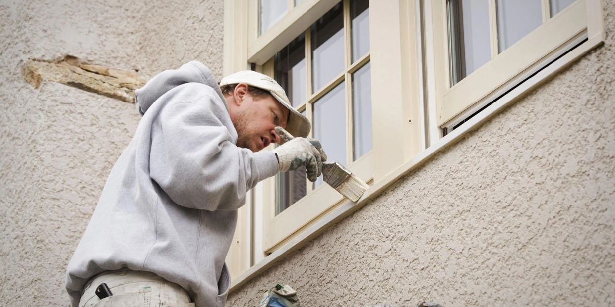 A man painting the trim around a window on a stucco wall.