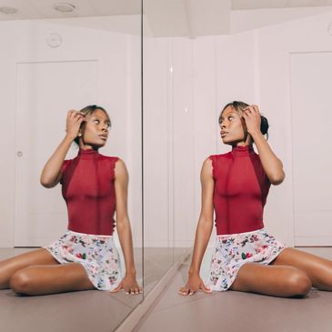 A woman in a red top and floral skirt sits, looking at her reflection in a mirror.