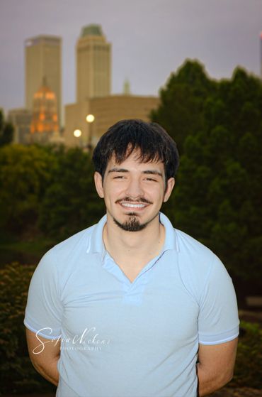 A young man smiling outdoors with city buildings in the background.