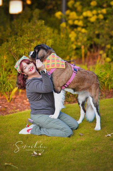 Woman kneeling on grass hugging a large dog wearing a colorful bandana.