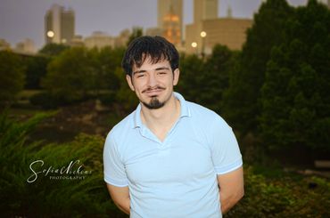 Young man in a light blue polo shirt smiling outdoors at dusk.
