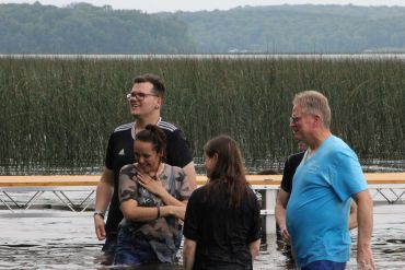 Four people standing in water near a dock with reeds and hills in the background.