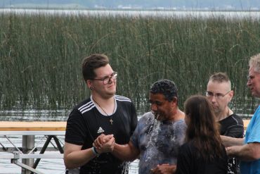A man is being baptized outdoors near a lake with others supporting him.