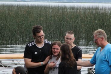 Group comforting a crying woman outdoors near water and reeds.