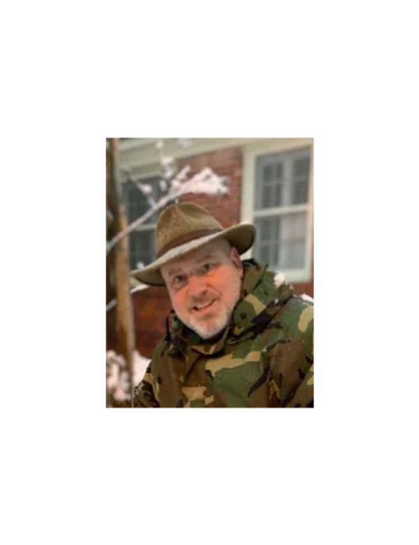 Man in camouflage jacket and brown hat smiling outdoors in winter.