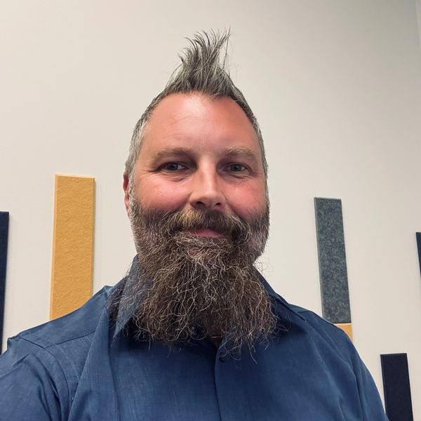 Man with a spiked mohawk and long beard smiling indoors.