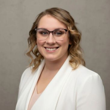 Woman with glasses and curly hair wearing a white blazer smiles.