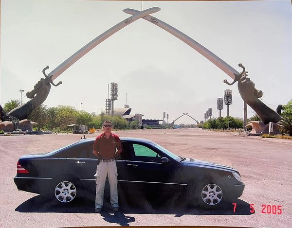 Man standing by a black car near large crossed swords monument.