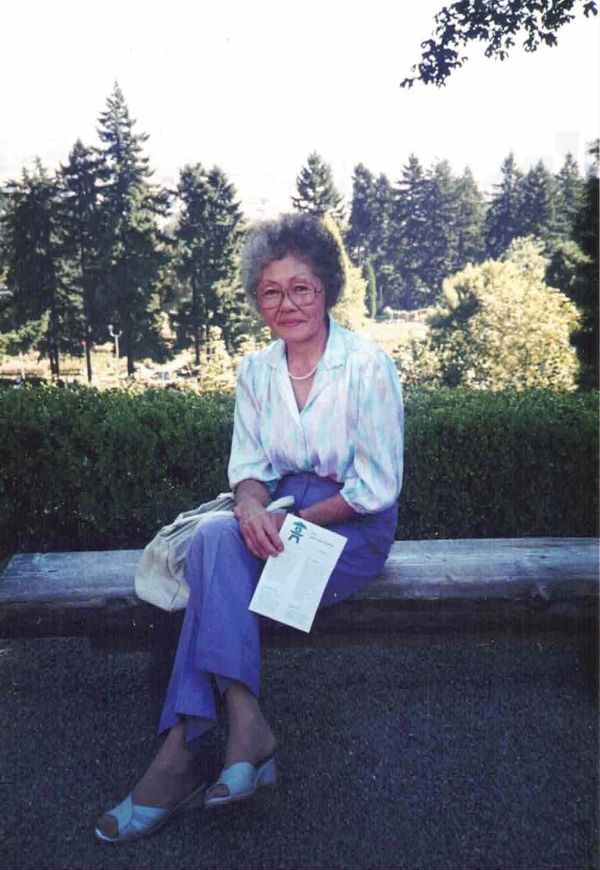 Elderly woman sitting on a bench holding a pamphlet in a garden.