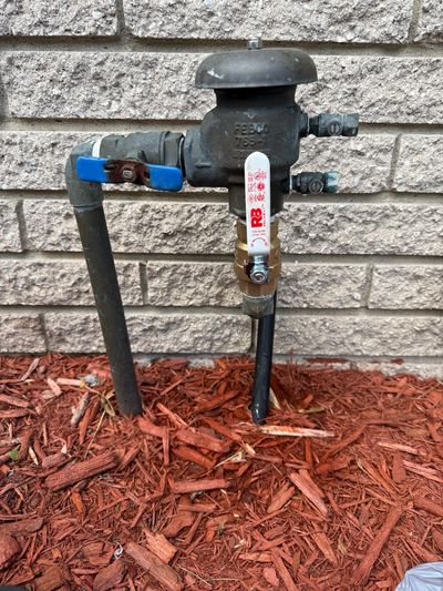 Outdoor water meter with valves against a brick wall and mulch bed.